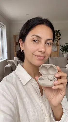 An actor in a bright living room holds an open fabric earbud case up to the camera while she talks, an ad for wireless earbuds.